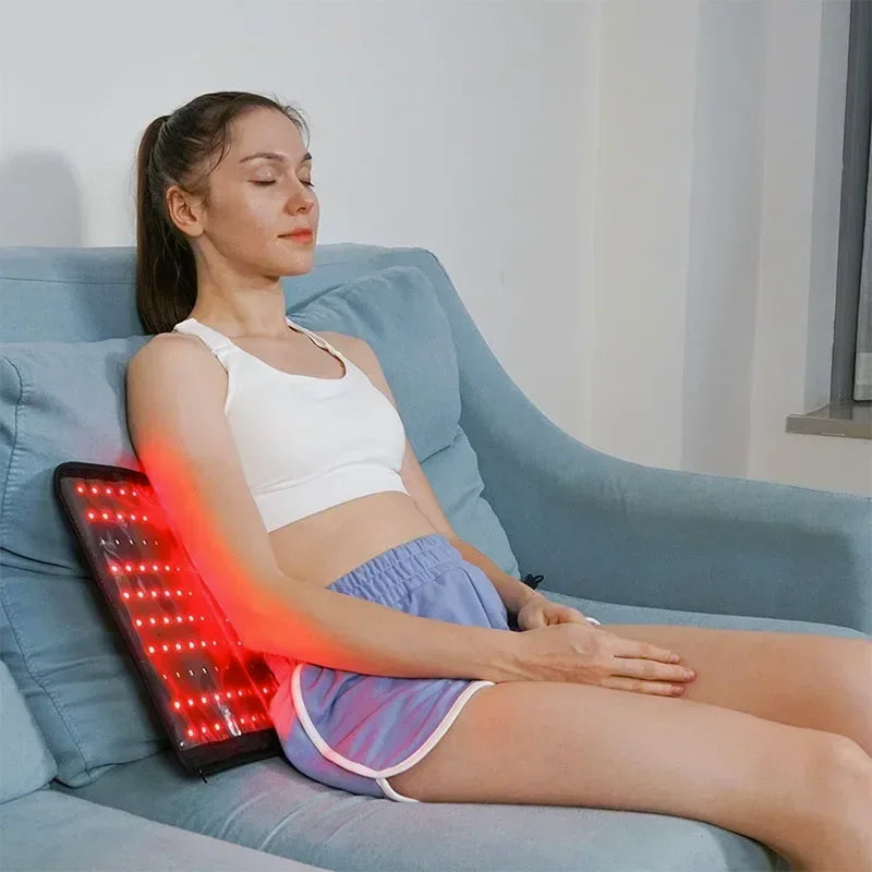 Woman using red light therapy pad on lower back while relaxing on a sofa